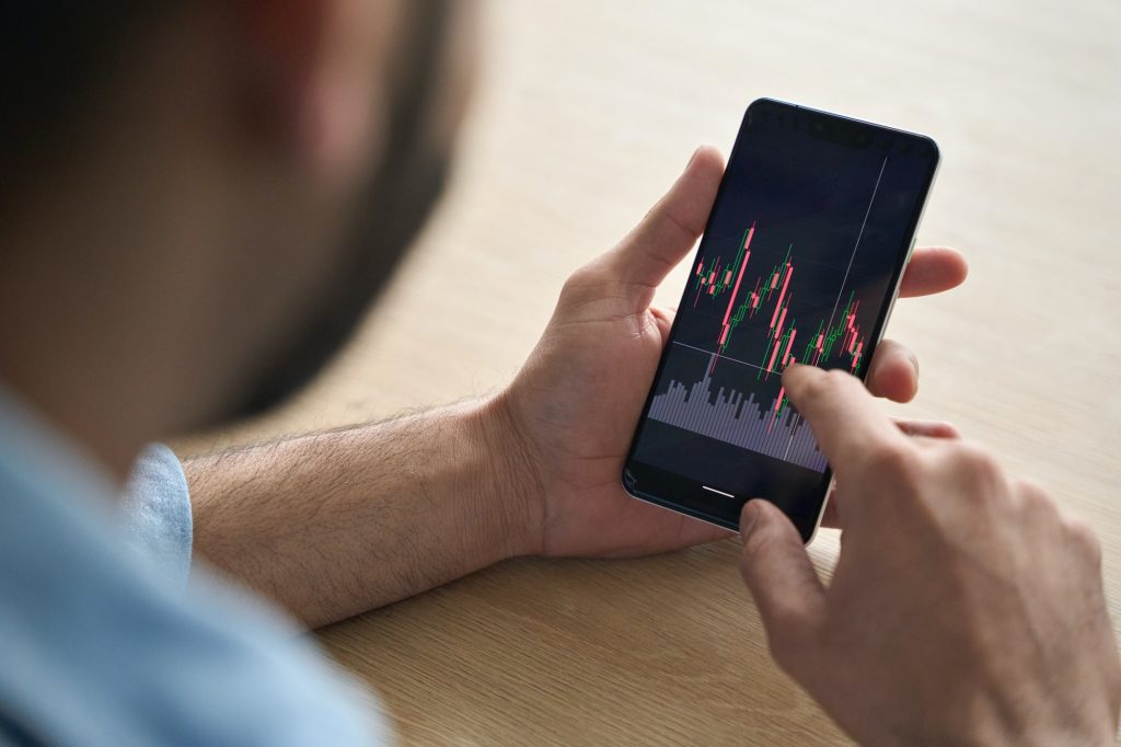 Closeup broker’s hands holding cellphone with stockmarket and graphs.