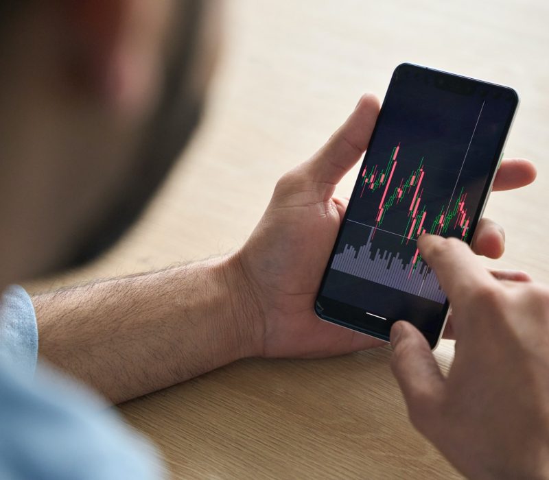 Closeup broker’s hands holding cellphone with stockmarket and graphs.