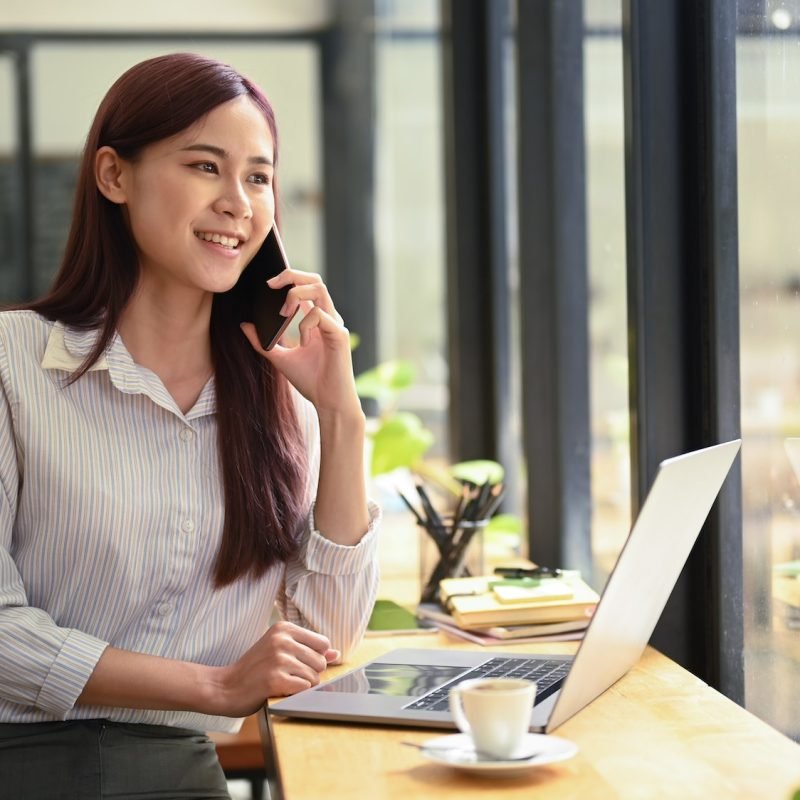 Successful young asian woman investor having phone conversation and using laptop at coffee shop.